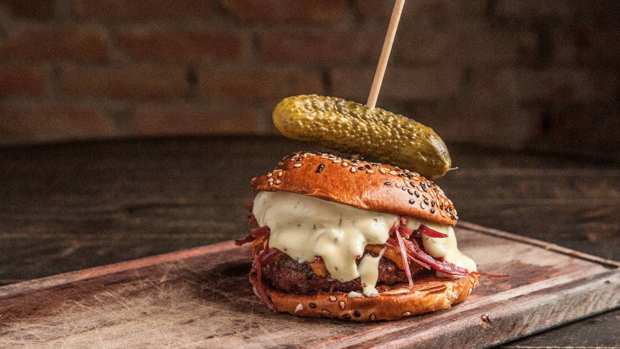 Foto de hambúrguer com pastrami e maionese escorrendo mais um picles de pepino inteiro por cima sobre tábua de madeira.