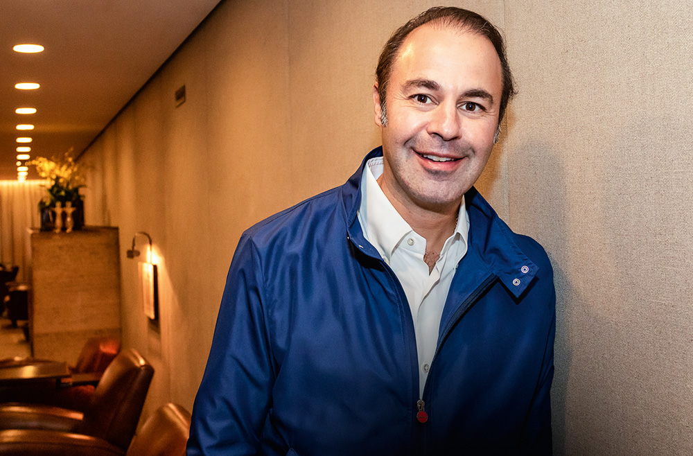 Retrato de Marcelo Pires, dos cotovelos para cima. Ele é um homem branco, calvo, de olhos e cabelos escuros. Usa uma camisa branca e uma jaqueta azul por cima.