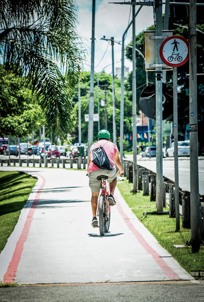 ciclovia radial leste Imagem mostra ciclista de camiseta vermelha andando em ciclovia cinza, entre um gramado e uma avenida.