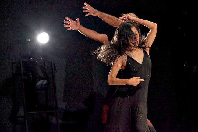 Sem Palavras_ Foto de Nana Moraes (46).jpg Imagem mostra mulher de vestido preto dançando . Ao fundo, um foco de luz branco.