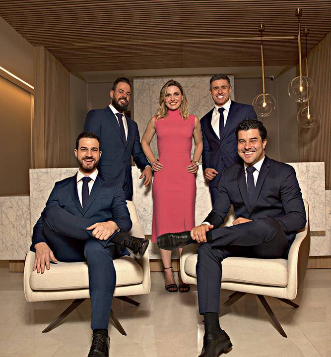 Cinco pessoas, 4 homens de terno e uma mulher de vestido midi rosa, posam para a foto. Dois homens estão sentados em poltronas bege na frente. As outras três pessoas estão atráz, com a mulher no meio. Todos são brancos e sorriem para a foto.
