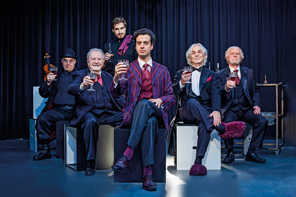 Imagem mostra 5 homens sentados, de terno, erguendo taças de vinho. Um homem em pé, ao fundo, veste uma roupa preta.