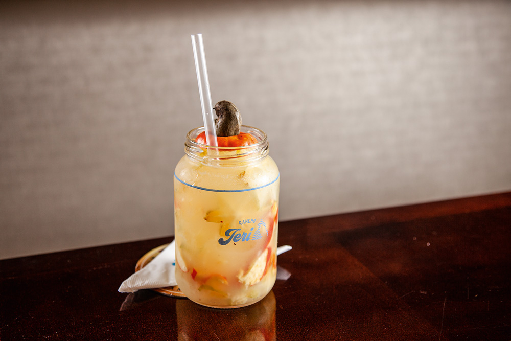 Rancho Jeri Caipirinha de cor levemente amarelada servida em pote de vidro decorado por parte de um caju