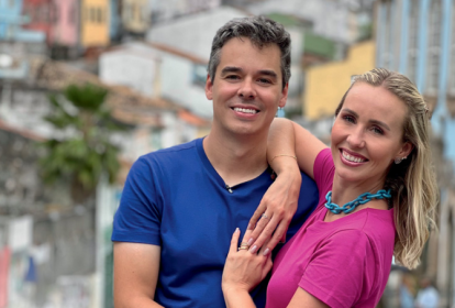 Imagem mostra um homem de camiseta azul e uma mulher de roupa rosa abraçados, sorrindo para a câmera.