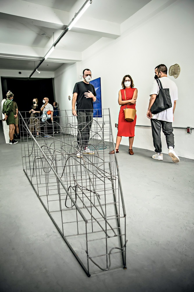São Paulo 12 fevereiro 2022Foto Alexandre Battibugli VSP Em um galeria, de paredes brancas e chão cinza, dois homens e uma mulher de vestido vermelho aparecem em destaque. À frente, uma escultura de arames está posta no chão.