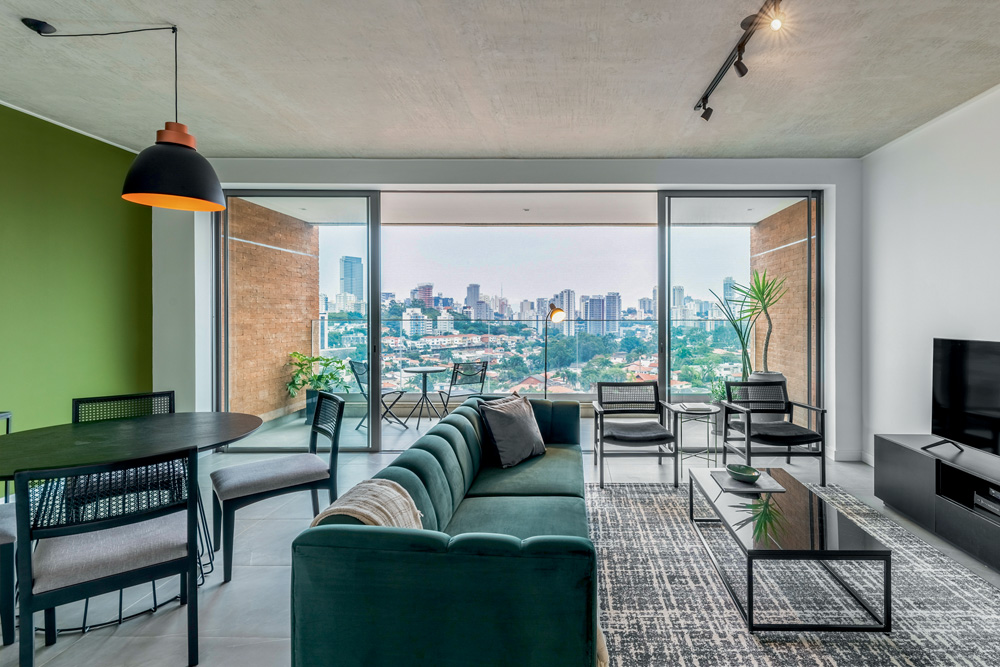 Guilherme Pucci(2).jpg Imagem mostra sala com sofá verde e cadeiras pretas. Ao fundo, uma paisagem com prédios e áreas verdes.