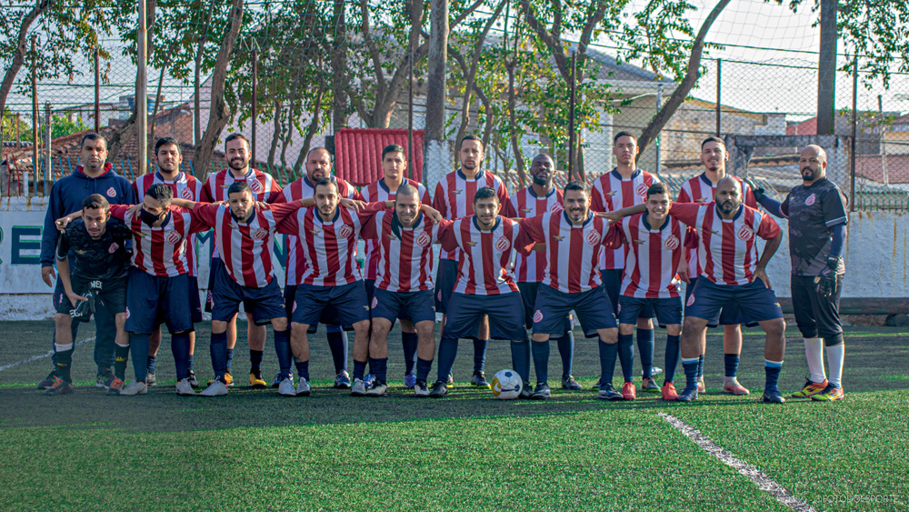 IMG_1653.jpg Imagem mostra time completo de futebol, com uniforme listrado vermelho e branco, enfileirados e abraçados.