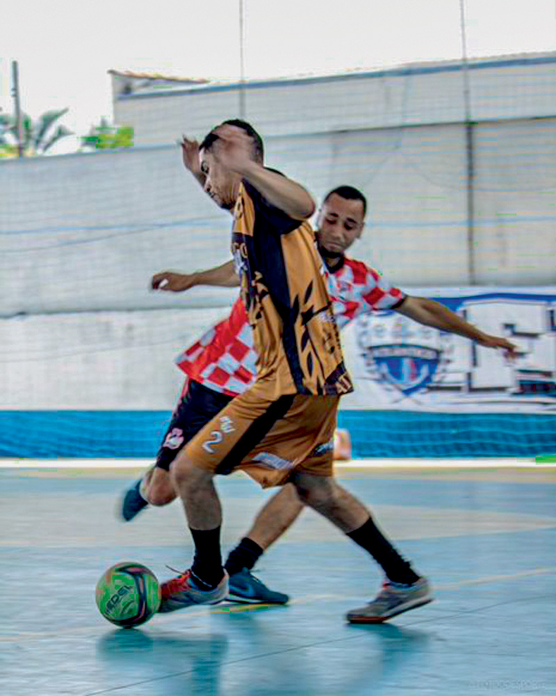 Liga da Quebrada Imagem mostra dois jogadores de futsal em quadra, um com uniforme amarelo e preto e outro com uniforme vermelho e branco. O de camiseta amarela está guiando a bola.