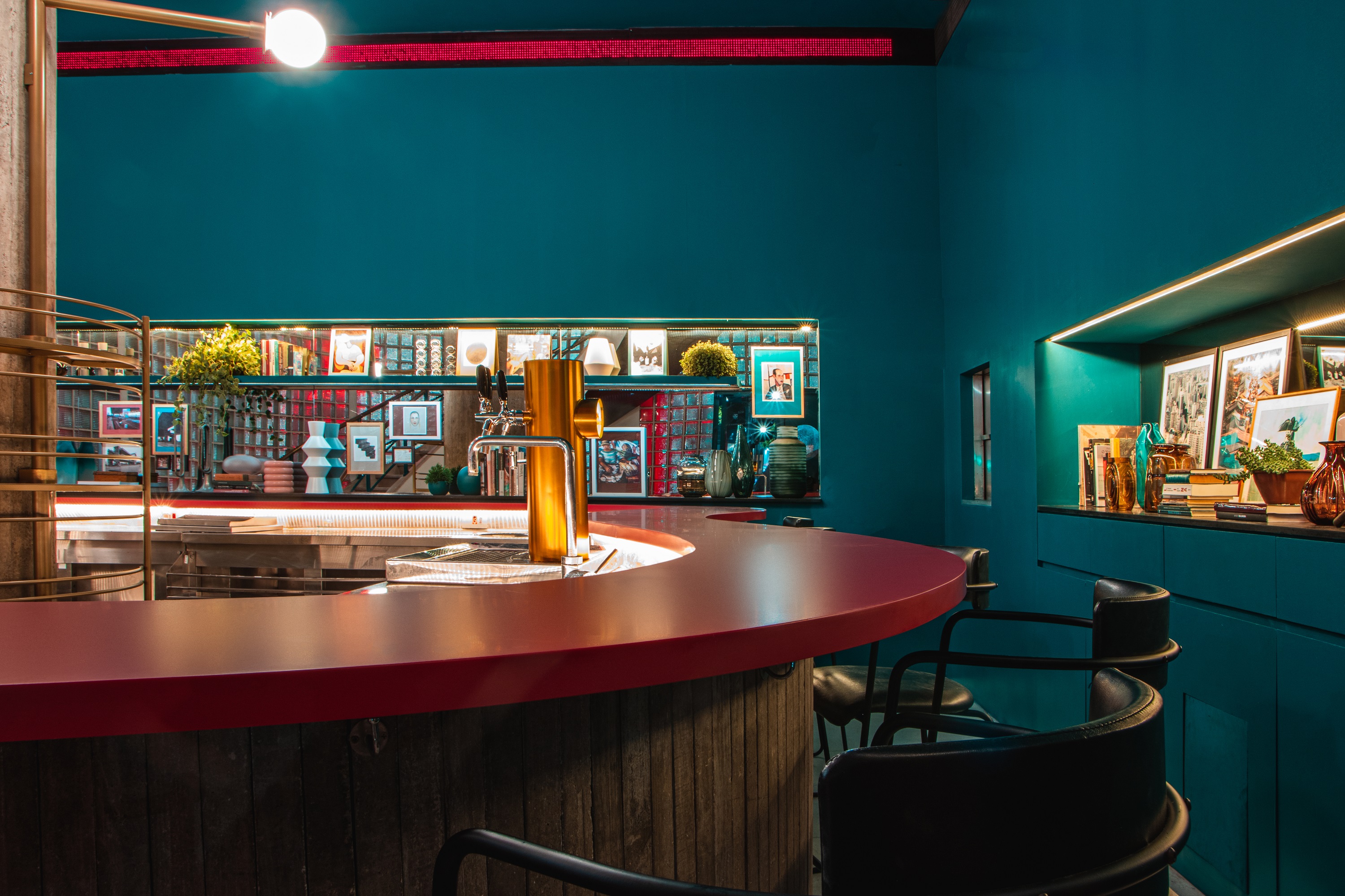 Ambiente de bar decorado por um balc&atilde;o de cor vermelha, banquetas altas e de cor preta e paredes de cor azul