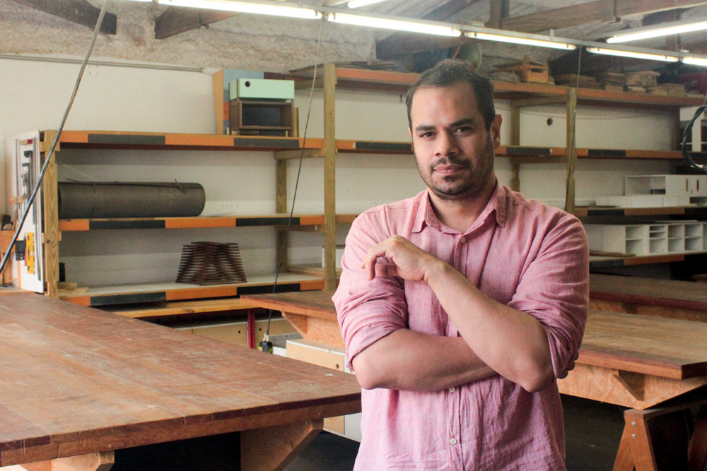 LAB74 (2 de 3).jpg Um homem branco, de cabelo e barba pretos, veste uma camisa rosa e posa em frente a prateleiras de madeira. O ambiente é uma marcenaria.