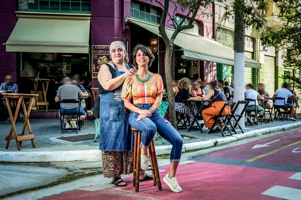 LASKARINA_9.JPG Duas mulheres, donas do empreendimento, posam na frente de um bar na Barra Funda. O local está movimentado. Ambas são brancas e usam jeans.