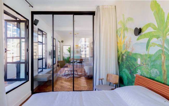 Latin Exclusive.jpg Imagem mostra quarto de casal com janelas de vidro e uma parede com um desenho de uma floresta à direita.