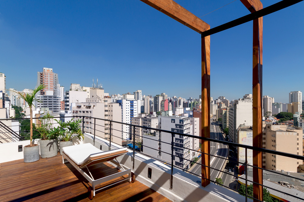 Latin Exclusive(1).jpg Imagem mostra terraço com chão de madeira e cadeira de praia. Um horizonte de prédios sob um céu azul preenche a imagem.