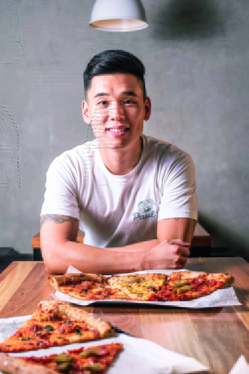 Paul’s Boutique Paul Cho posa de braços cruzados sobre mesa onde estão dispostos diferentes pedaços de pizza