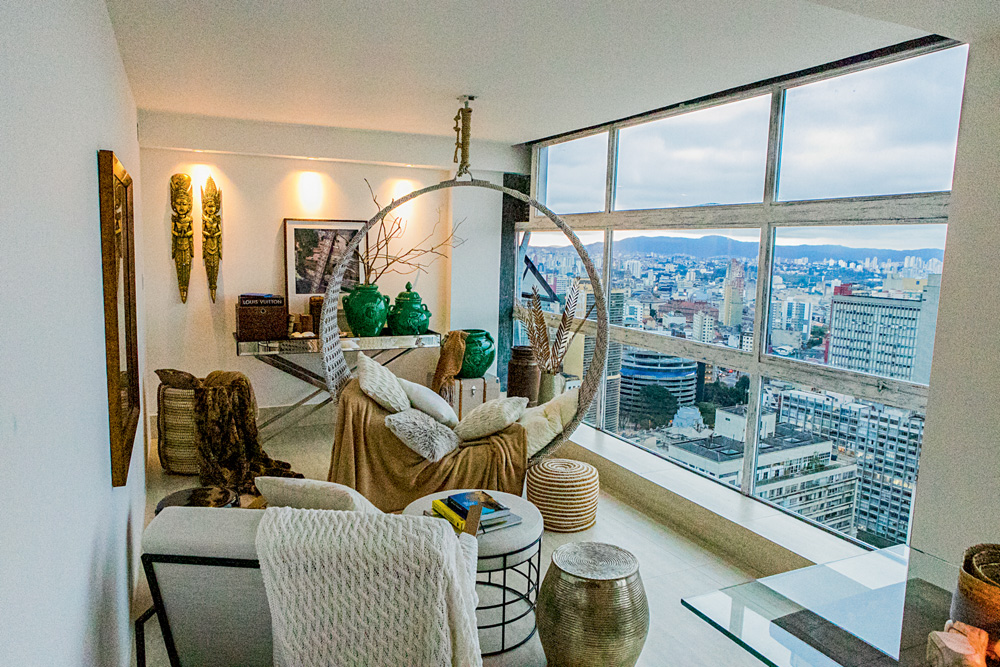 Apartamento Varandas Sidnei Tavares.jpg Imagem mostra sala com diversos móveis, paredes brancas e uma janela grande de onde é possível ver um horizonte de prédios com um morro ao fundo.