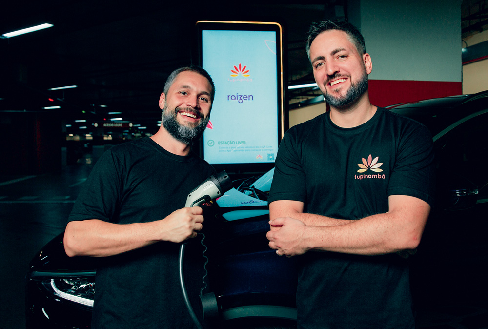 tupi investimento horizontal Imagem mostra dois homens de camisetas pretas, sorrindo e segurando plug de abastecimento de carros elétricos.