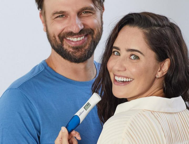 Imagem mostra casal sorrindo, o homem de camiseta azul e a mulher de roupa branca. Ela segura um teste de gravidez na mão.