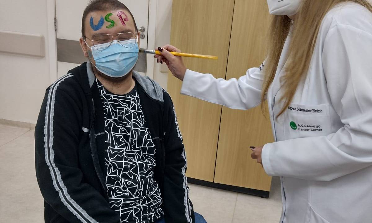Imagem mostra Jovem sentado em uma cadeira e uma mulher e pé pintando o seu rosto
