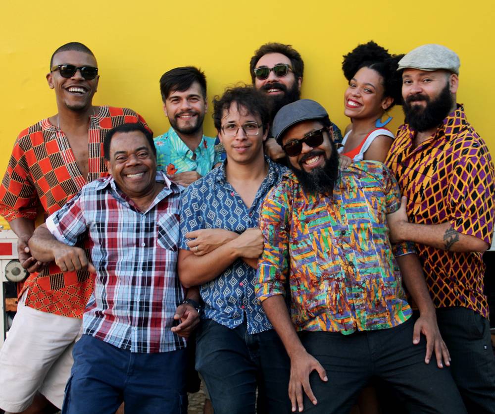 crédito Tarsila Danieletto.jpg Sete homens e uma mulher vestem roupas coloridas e posam juntos sorrindo em frente a uma parede amarela. São um grupo musical.