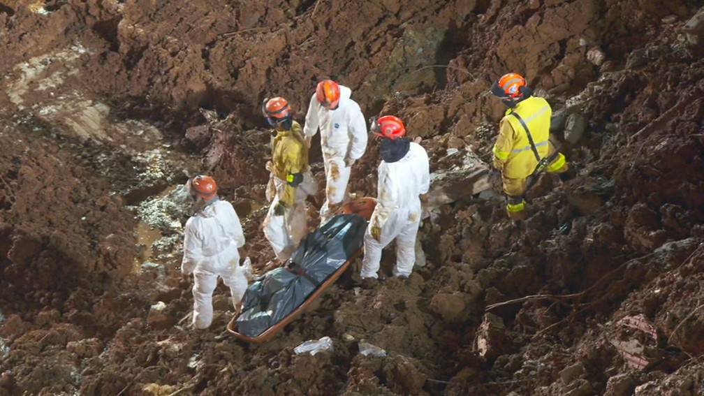 A foto mostra trabalho do corpo de bombeiros em área atingida pelas chuvas. Eles carregam uma maca com um corpo encontrado nos destroços