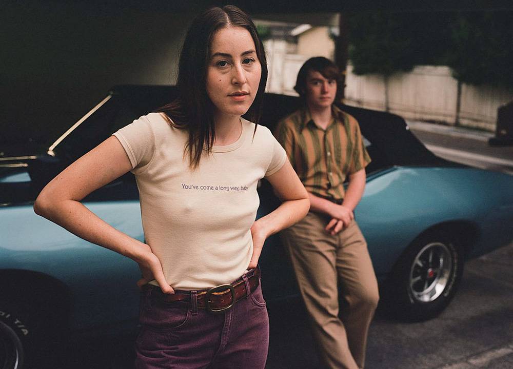 Imagem mostra mulher, de camiseta branca e calça roxa, à frente de carro azul. Um homem de camisa listrada está apoiado no carro, atrás dela.