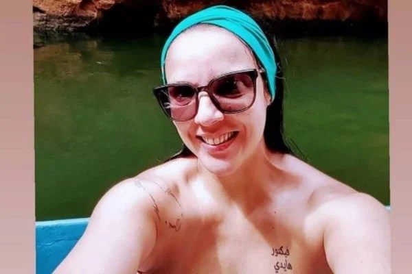 Imagem mostra Mara, de óculos e com faixa no cabelo, sorrindo em selfie, com rio atrás