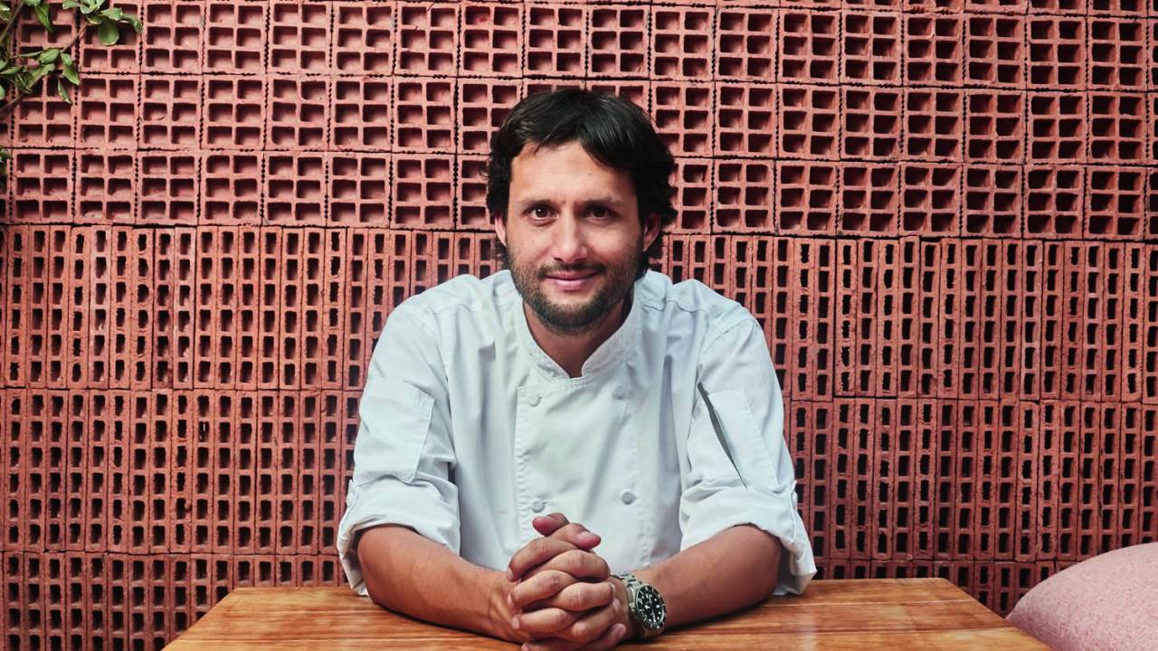 Jaime Pesaque posa sentado e de braços apoiados sobre uma mesa de madeira frente a uma parede de tijolos