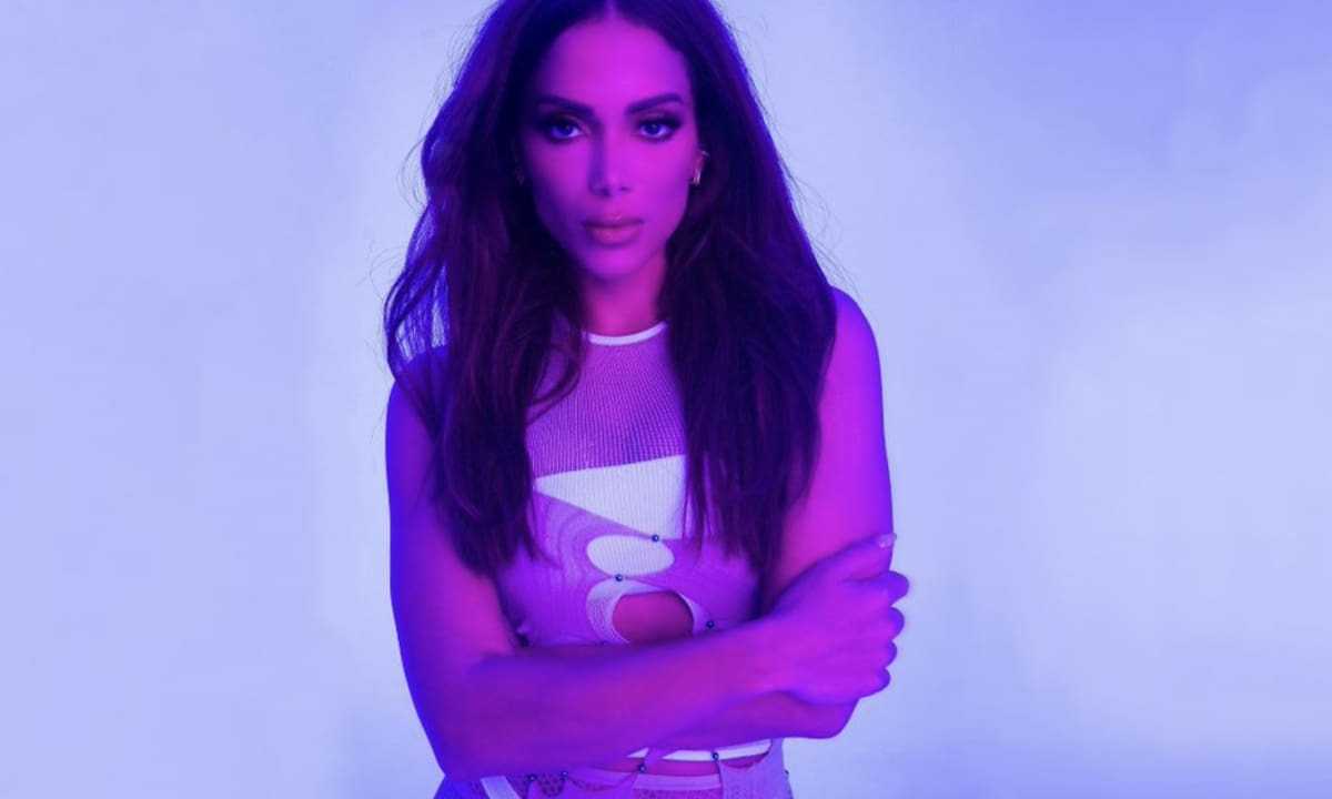 Anitta é uma mulher branca, de cabelos lisos e escuros. Usa um body branco e está sob uma luz roxa em seu clipe