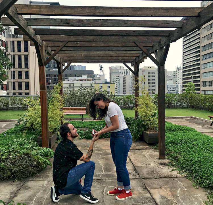 Felipe, um homem branco, de cabelos lisos e castanhos, ajoelha e pede Beatriz em casamento em um jardim suspenso. Ela também é branca, tem cabelos castanhos e cacheados. Ambos usam caça jeans e camiseta