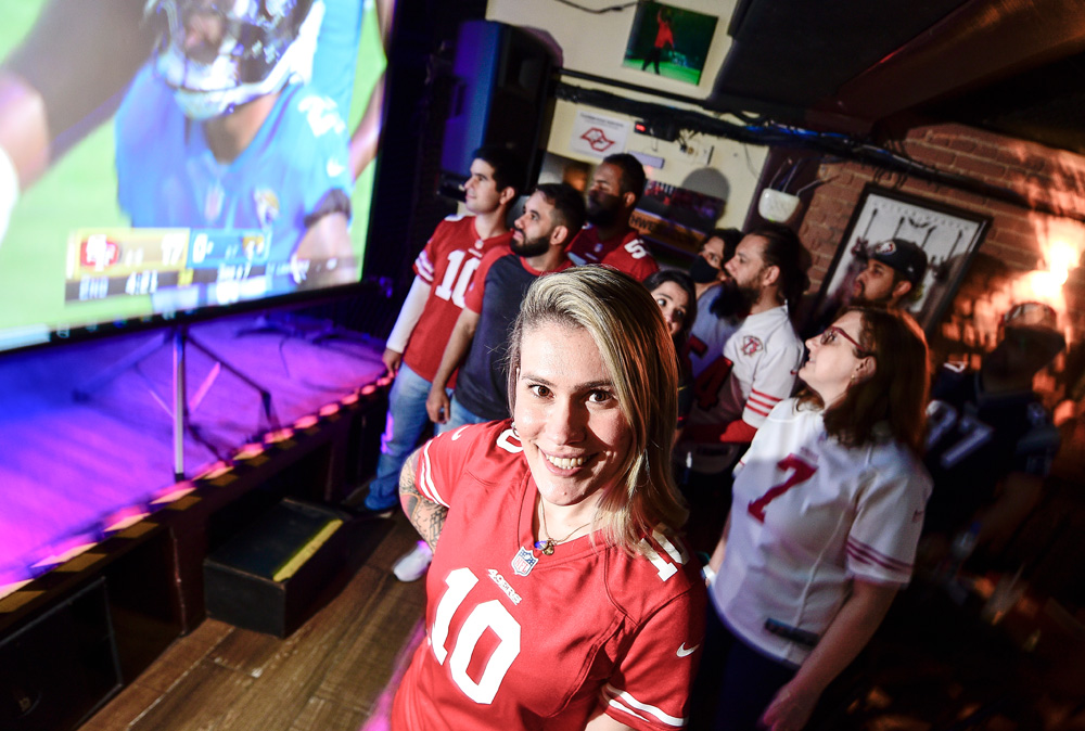 LAIS_5 Imagem mostra mulher loira vestindo camiseta de time vermelha em meio a aglomeração de pessoas em bar.