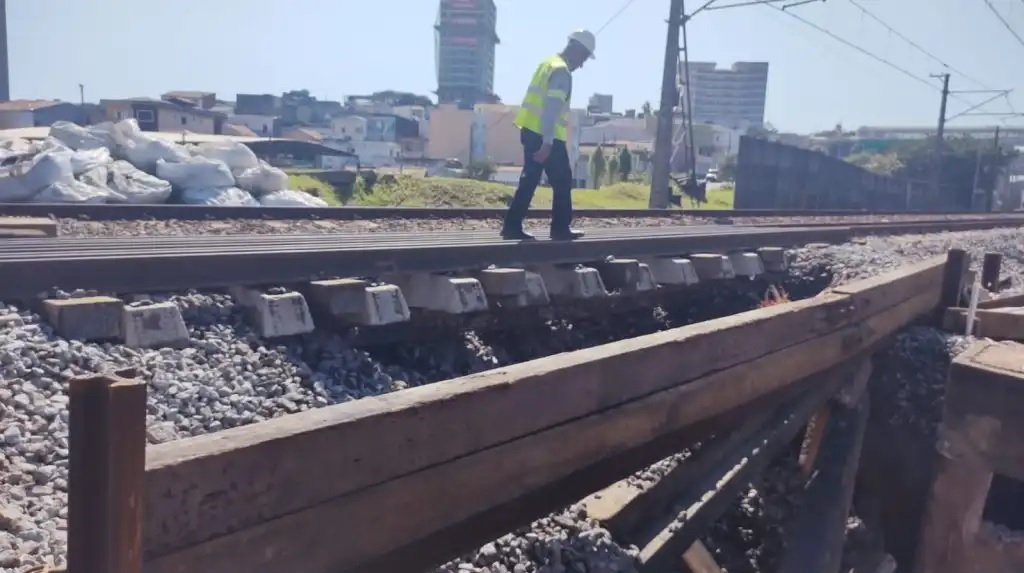 Foto exibe trecho da Linha 11-Coral com trilhos em foco e funcionário com capacete de segurança ao fundo.