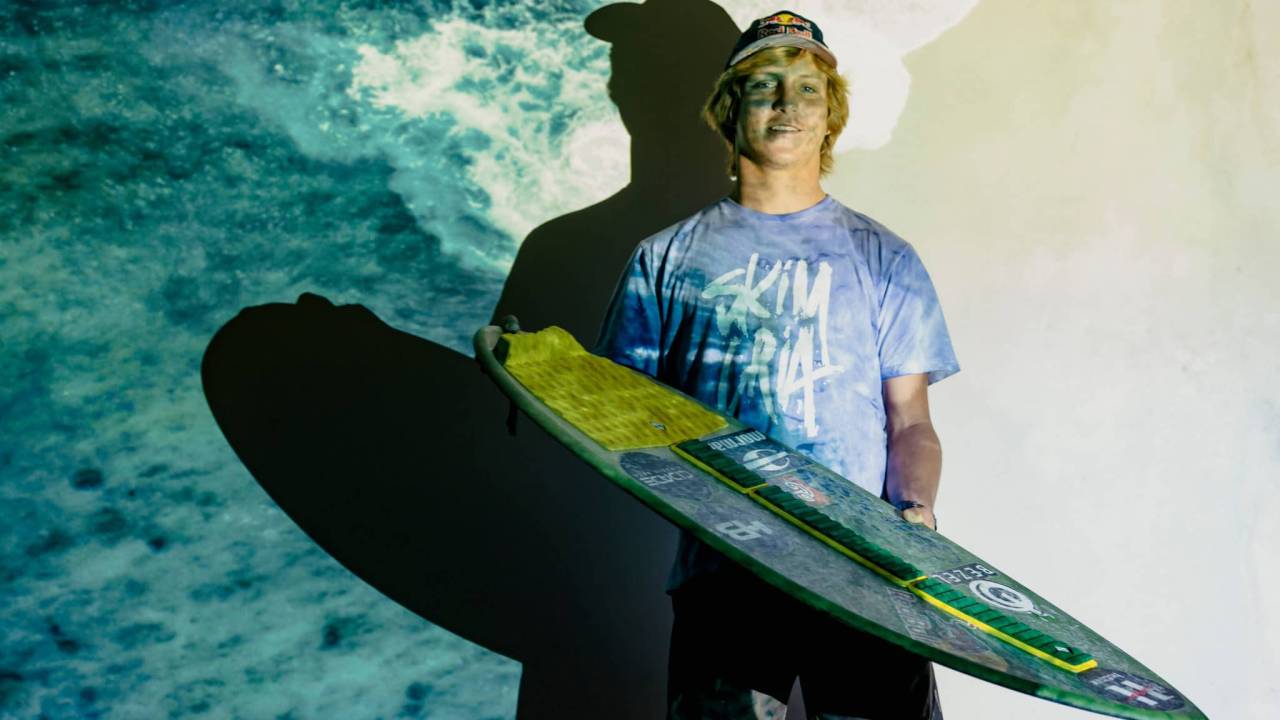 Lucas Fink posa segurando prancha verde de pé em fundo com imagem do mar.