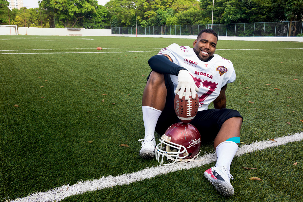 Futebol Americano Imagem mostra homem vestido de uniforme de futebol americano, segurando uma bola oval, sentado nda grama.