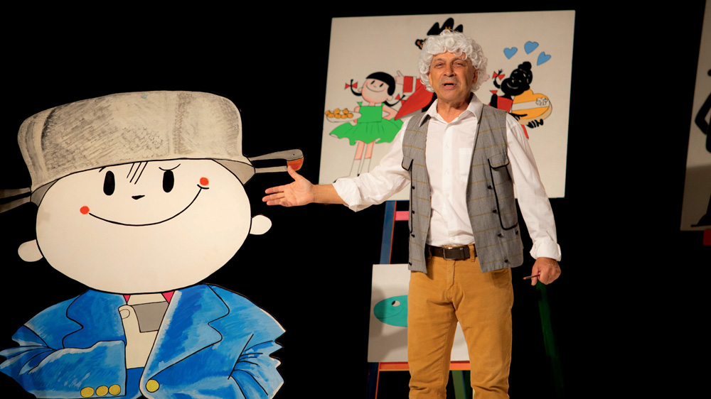 Um ator faz Ziraldo em cena. Ele está em seu ateliê e, ao lado, há um grande cartaz com o Menino Maluquinho