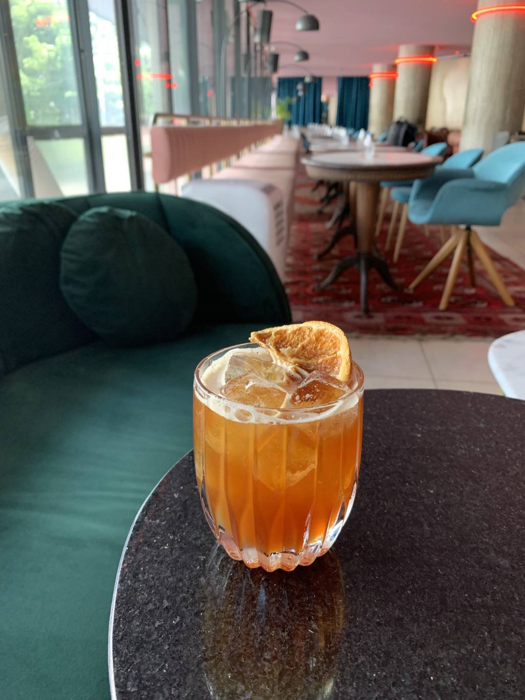 Riviera Bar Foto de drinque de cor alaranjada servido em copo baixo decorado por um pedaço de laranja desidratada