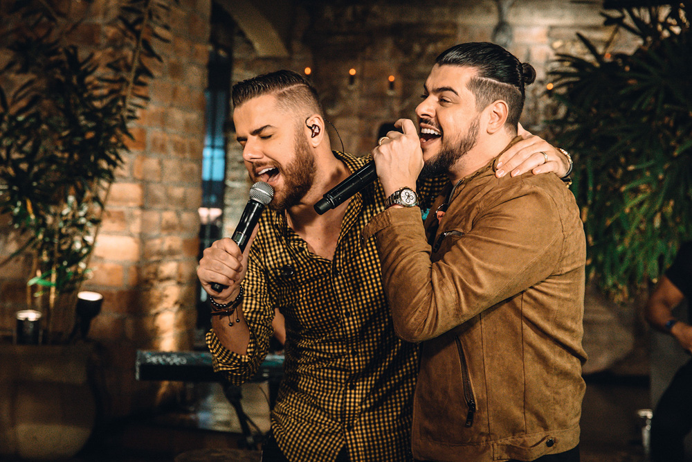 Imagem mostra dois homens abraçados cantando em dois microfones.