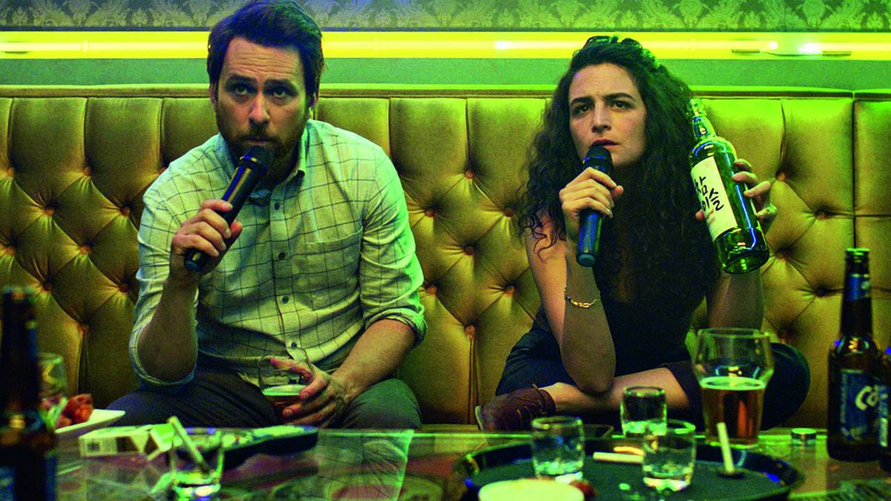 Imagem mostra um homem e uma mulher sentados em sofá, segurando microfones e tomando vinho.