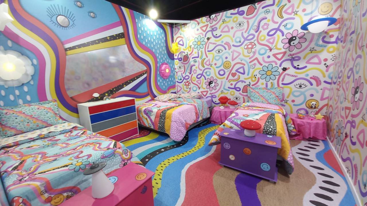 Quarto colorido em tons pastéis, com três camas, baús coloridos e mesas com abajures de cogumelo