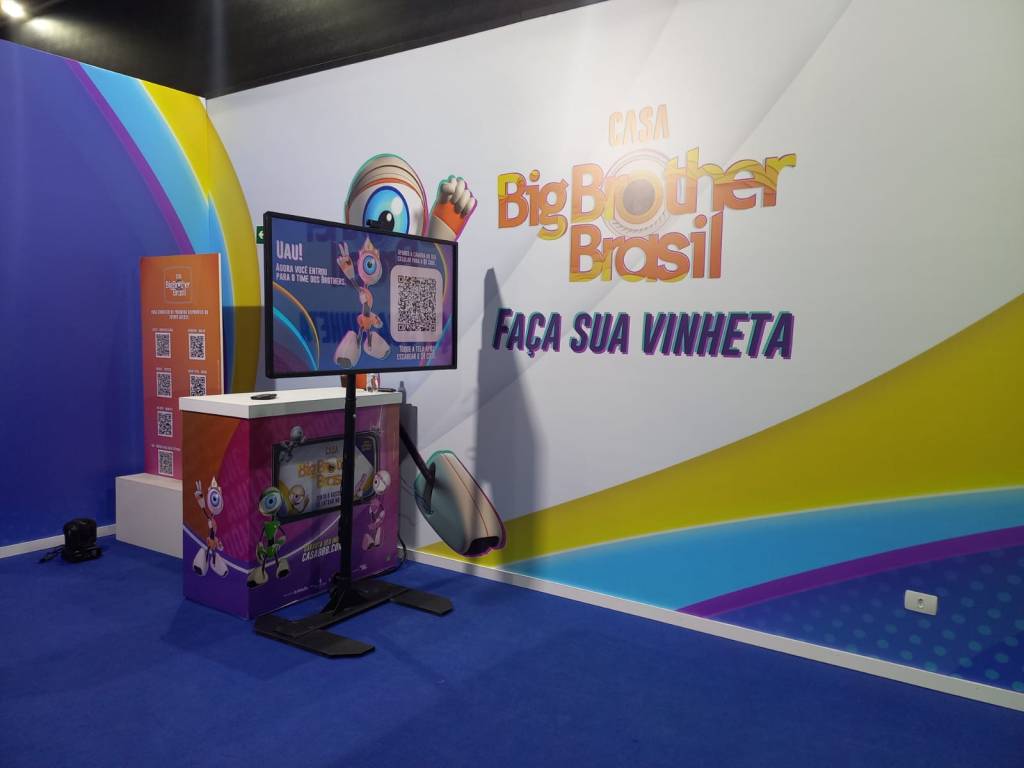 vinheta Uma parede ampla com o logo do BBB com um televisor suspenso ao lado. Chão é feito de carpete azul