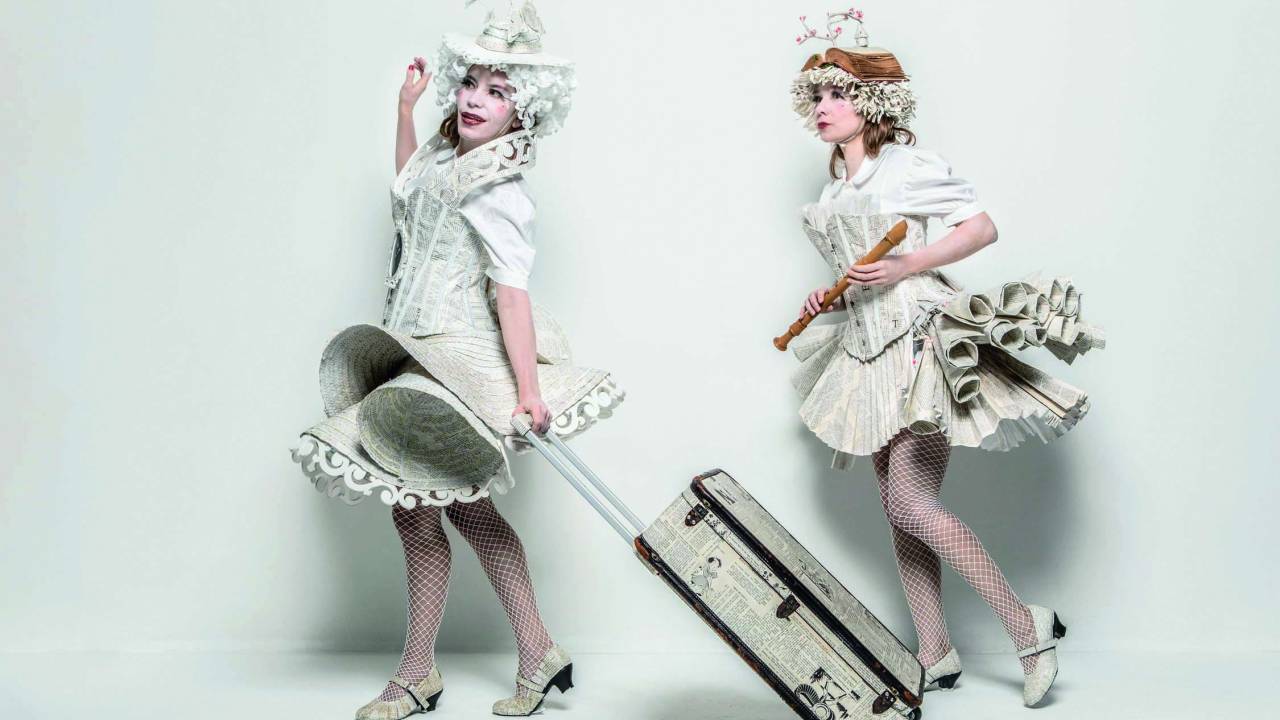 Duas mulheres vestem figurinos delicados, que lembram roupas da Era Vitoriana, feitos de páginas de livros. Ambas são brancas. Uma delas vai à frente e carrega uma mala de rodinhas, também revestida de páginas, e a outra vai atrás com uma flauta doce nas mãos
