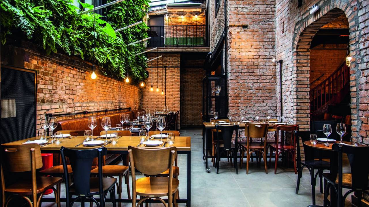 Imagem do salão do restaurante. Há mesas e cadeiras de madeira. Em uma das paredes, há uma decoração com plantas