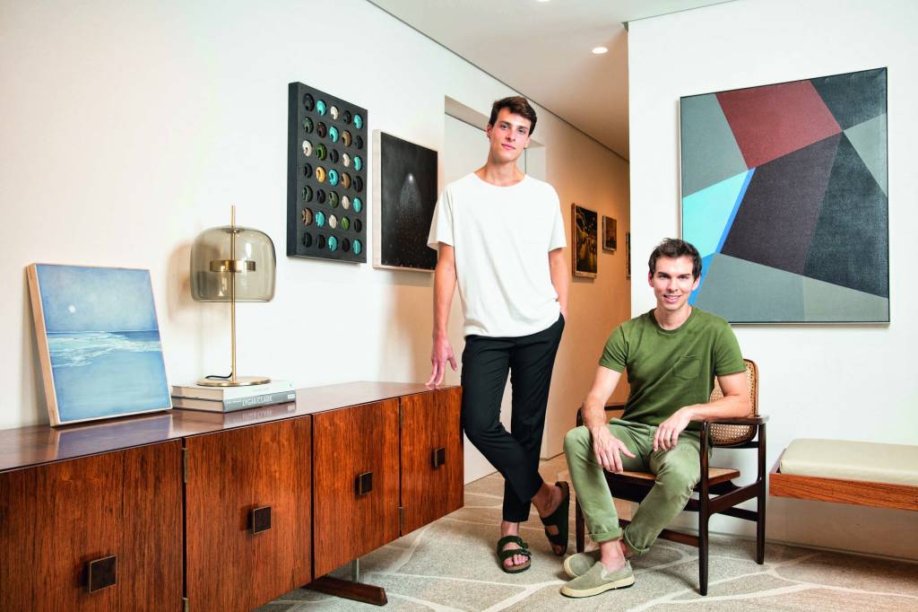 COLECIONADORES DE ARTE Imagem mostra dois homens, um em pé, de camiseta branca e calça preta, e o outro sentado, de camiseta e calça verdes. No ambiente, paredes brancas com pinturas penduradas.