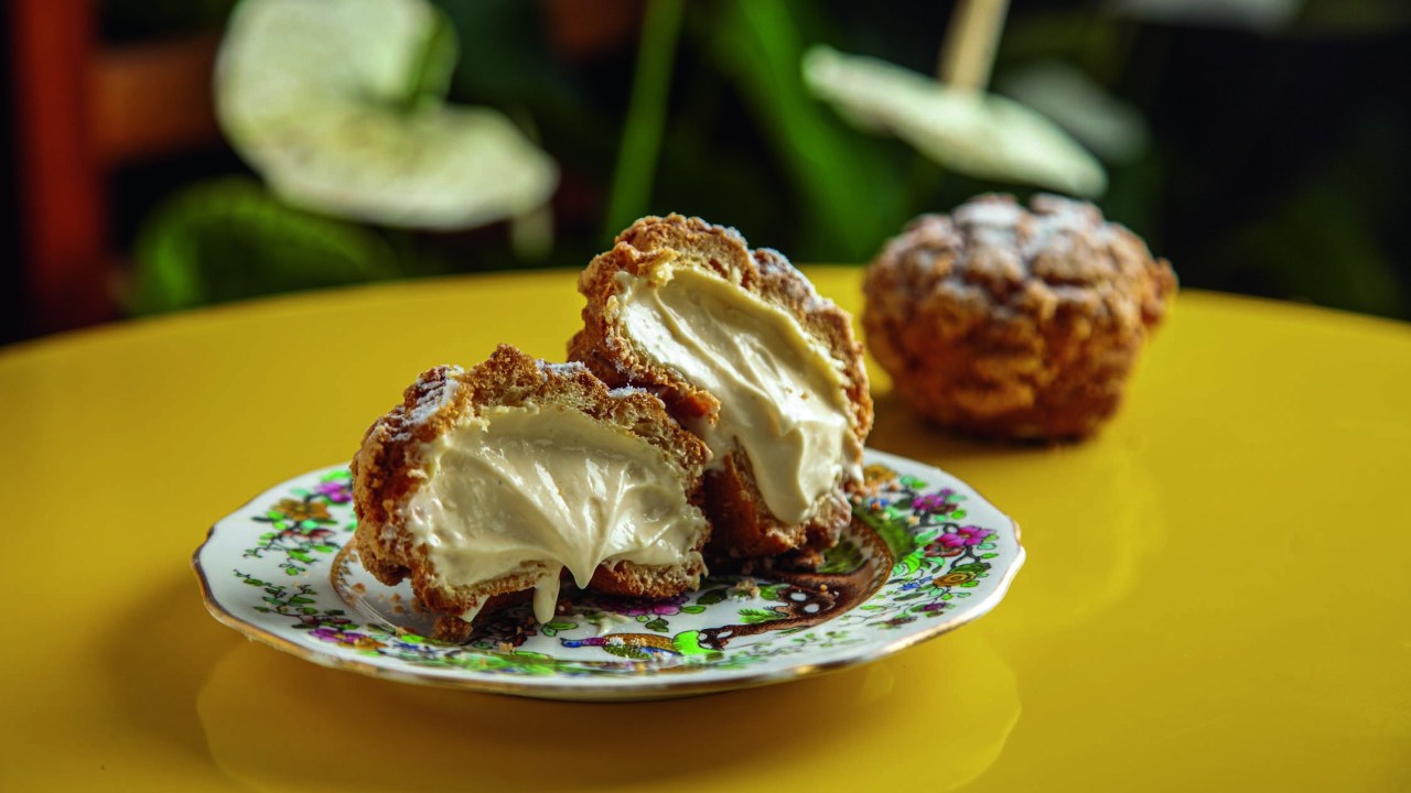 Carolina choux recheada por creme de cor clara cortado no meio e servido sobre prato de louça decorado sobre mesa de cor amarela
