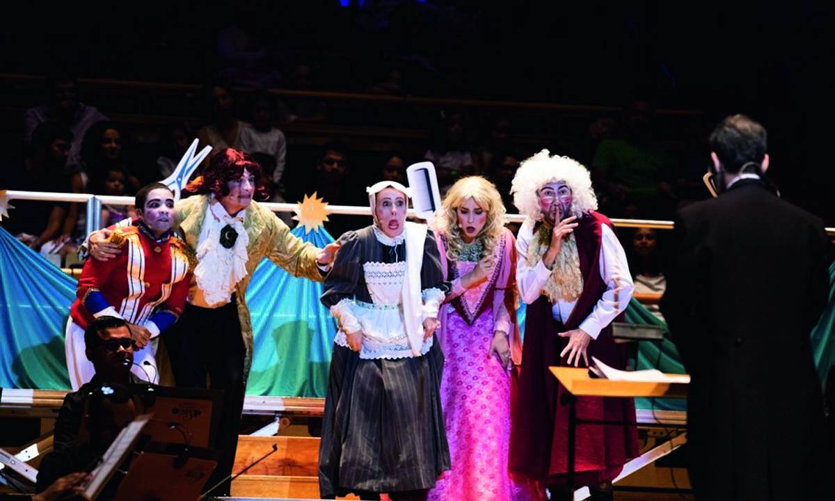 Cinco atores vestidos com figurinos que simulam roupas do século XIX em cima de um palco. É possível ver o maestro que rege a orquestra de costas, em frente aos atores