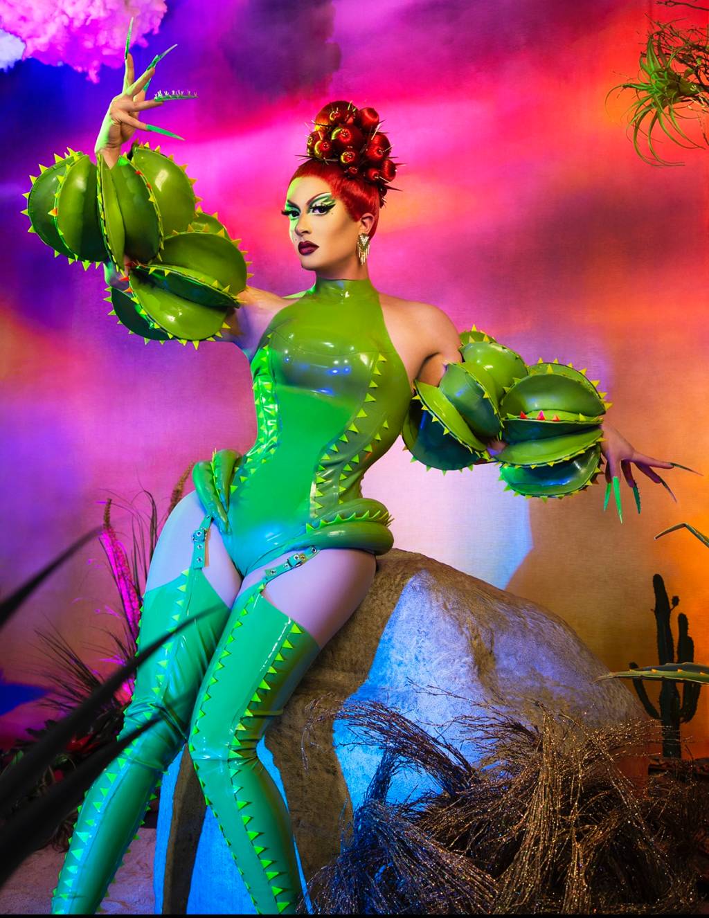 DenaliCactus Drag queen Denali posa em fundo psicodélico vermelho e veste vestido verde com peruca ruiva.