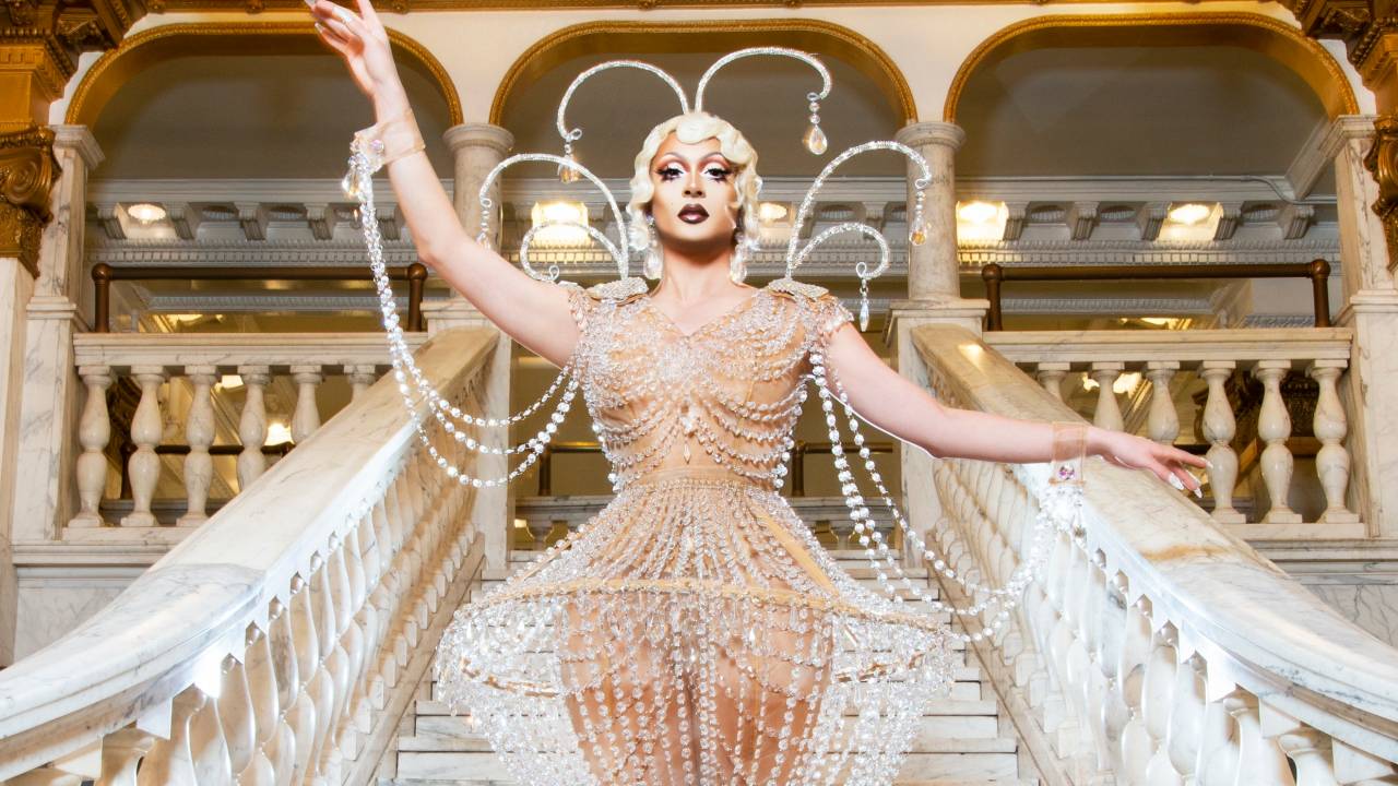 Drag queen Denali posa em escadaria branca com vestido de pedras, uma das mãos para cima, e o teto dourado em evidência.