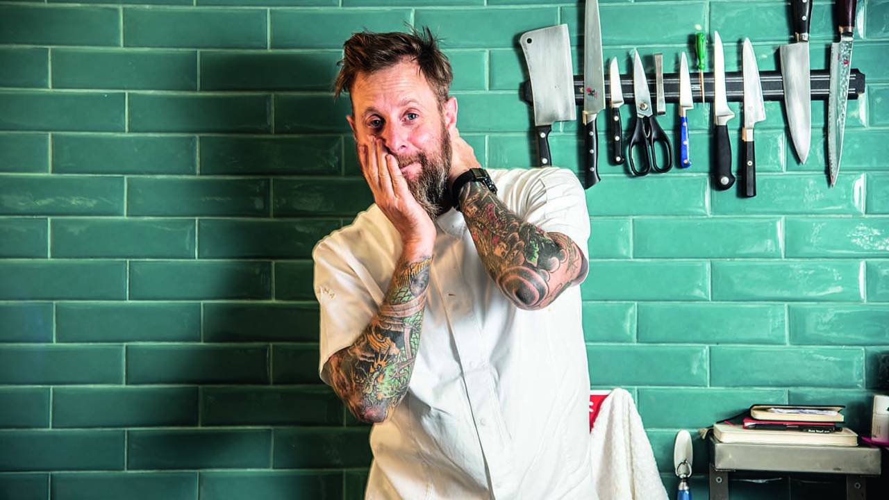 Chef André Mifano posa vestindo avental em frente à parede de azulejos de cor azul. Na mesma parede, há um faqueiro pendurado à direita de Mifano.