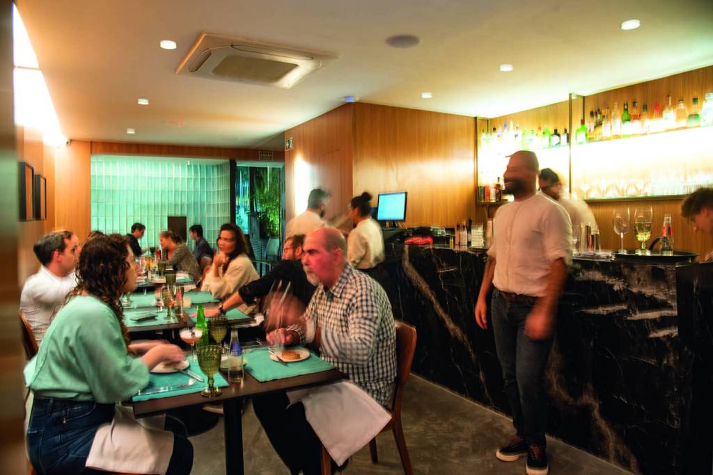 Donna Ambiente de restaurante iluminado por brancas. Mesas são dispostas lado a lado em frente à parede e, ao fundo, há um balcão de mármore escuro onde encontra-se o bar, decorado por garrafas expostas em prateleiras
