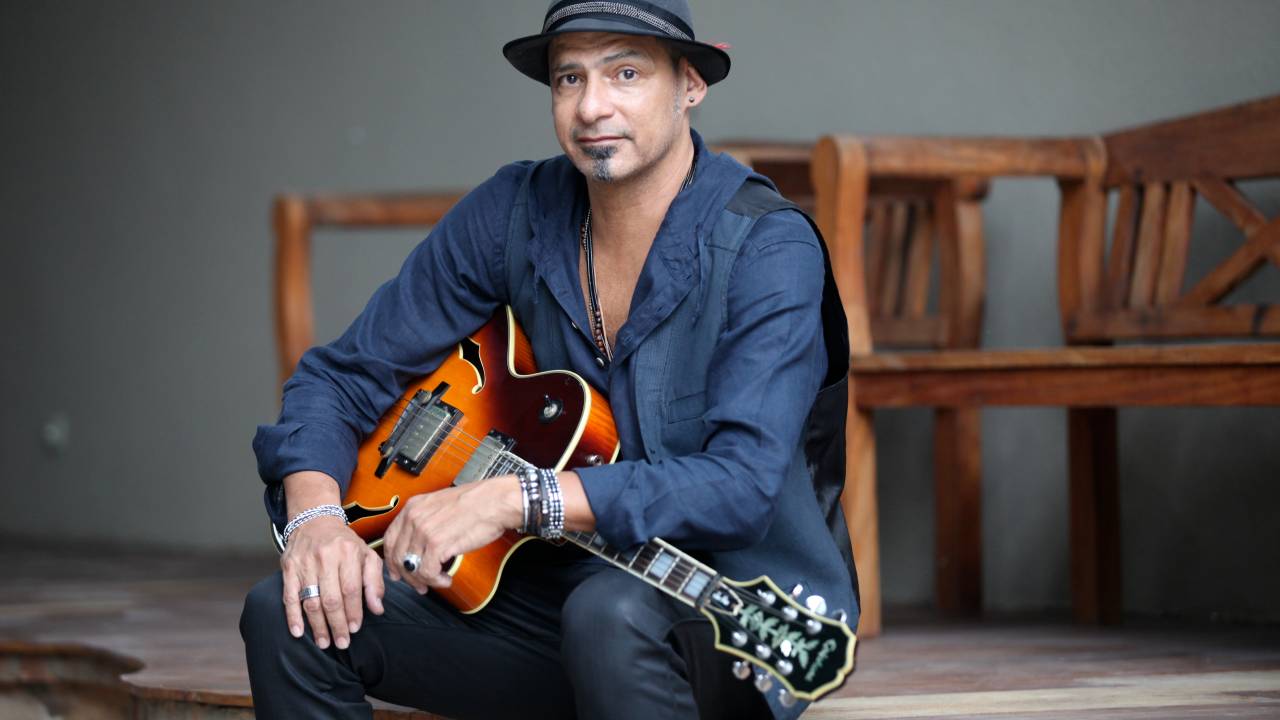 Imagem mostra homem de camisa e chapéu, segurando uma guitarra.