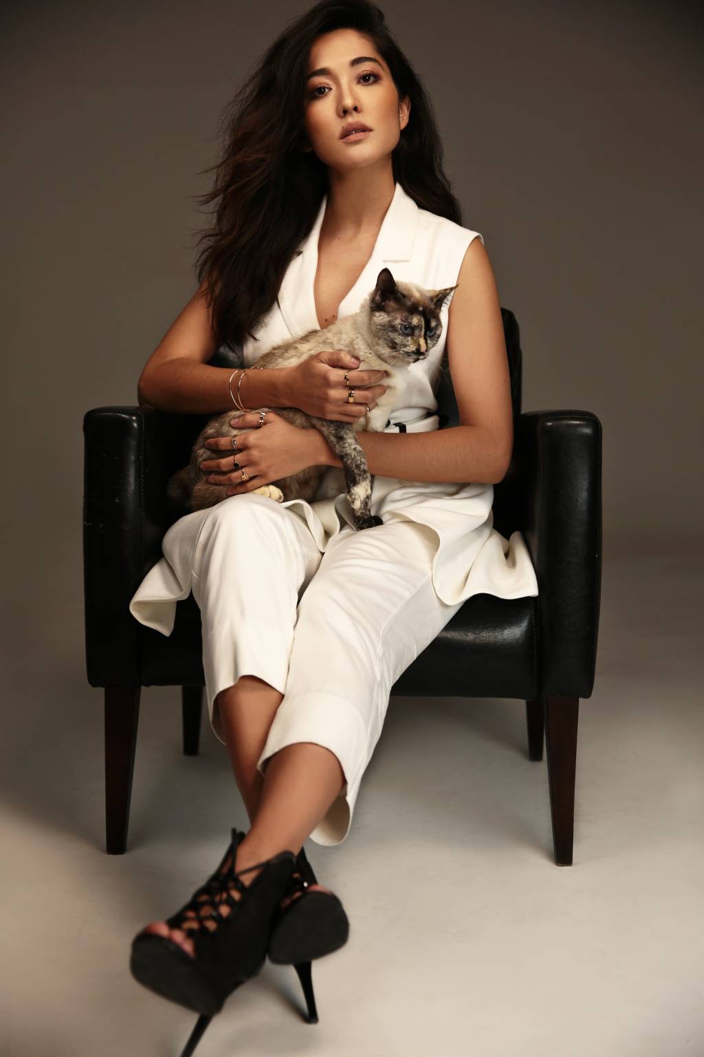 Jacqueline Sato (PUPIN + DELEU-DIVULGAÇÃO) Jacqueline Sato posa com gato no colo enquanto está sentada em uma poltrona preta. Ela veste roupa toda branca e salto alto preto. Encara a câmera com olhar sério.